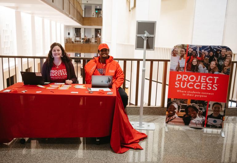 Two people wearing Project Success clothing sit smiling behind a red table on a balcony in Northrop. A Project Success banner stands next to them.