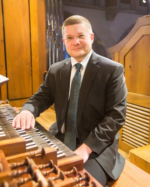 An organist with short, medium brown hair, oval glasses, wearing a dark grey suit paired with a grey and teal polka dot tie, sits on a wooden bench, resting their right arm on the keys of the organ instrument beside them. 