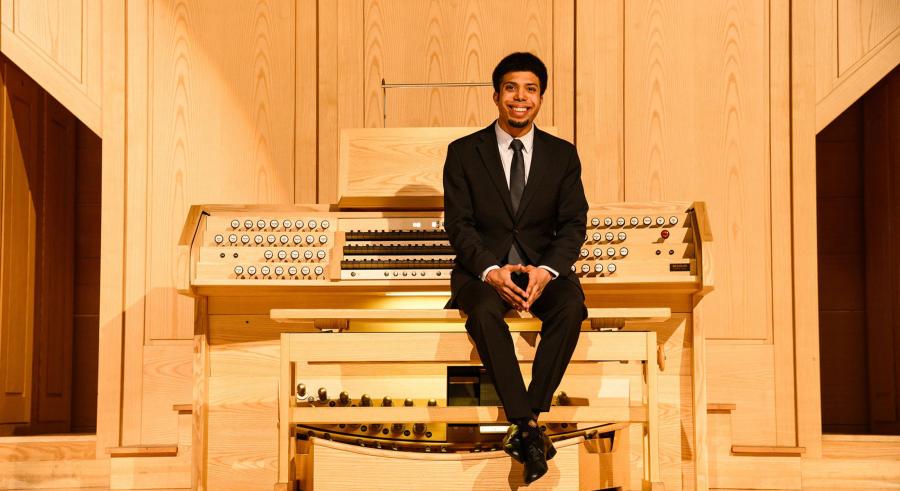 An organist with dark hair wearing a black suit and tie sits in a recital hall with their back towards a wooden organ console and fingers intertwined and placed on their knees.