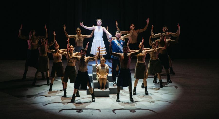 A group of dancers with black pants stand with their arms creating a U shape around a dancer wearing a white dress, a dancer wearing a militaristic suit, and a dancer in white sitting on steps.