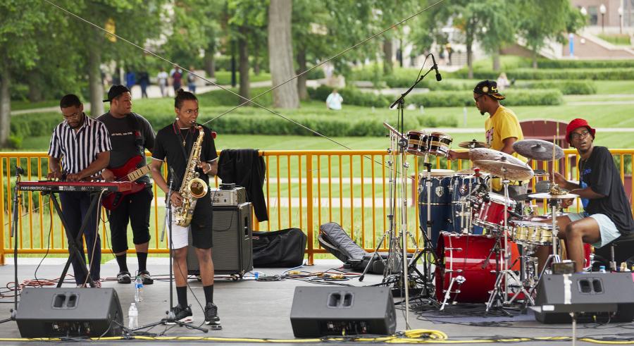 Five performers from LA Buckner perform music from their album, BiG HOMiE, on the Northrop Plaza Stage. They are playing the keyboard, guitar, saxophone, drumset, and congas.