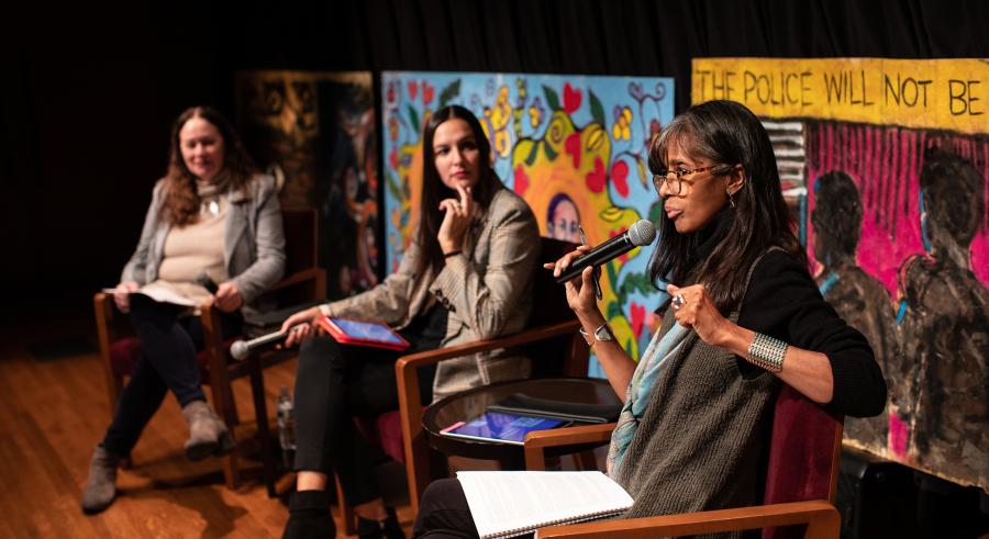 Three people sit onstage. The woman closest to the camera is holding a microphone and speaking while the other two listen. Behind them are three murals with various art styles.