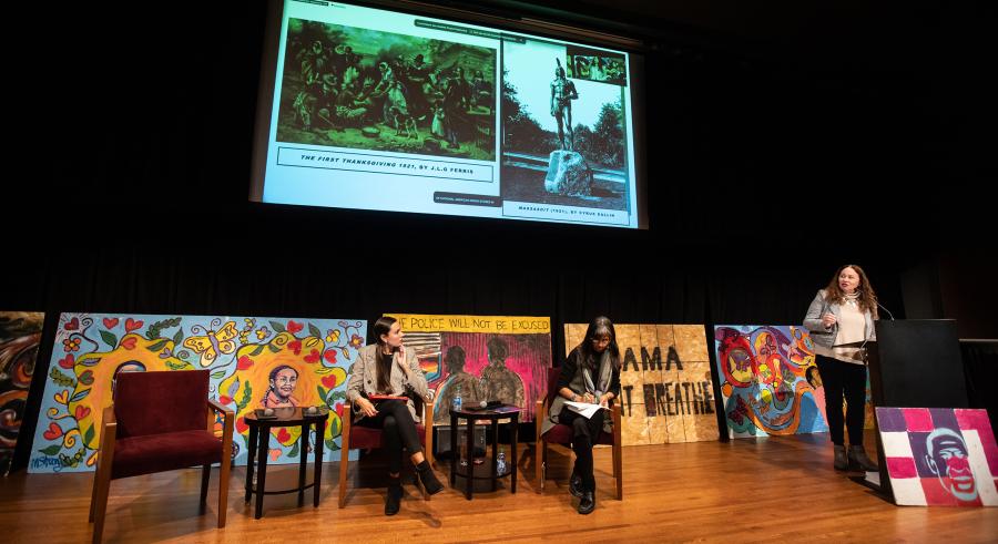 One person stands behind a podium, discussing photos of a monument and a mural on a large screen. The other two speakers sit in chairs in front of various murals, listening and writing.