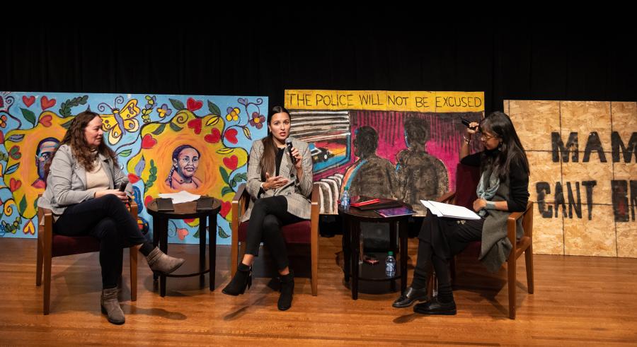 Three presenters sit on stage in front of various murals and a projected screen asking audience members to “Join the Conversation” and ask questions via an online platform called Slido.