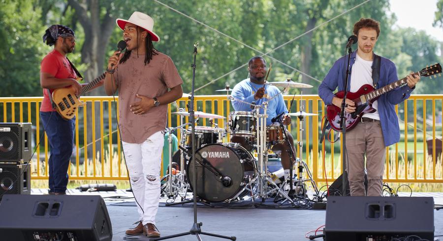 Four musicians making up DeTrell Melodies performing on the Northrop Plaza Stage. They are playing bass guitar, guitar, drums, and one is singing.