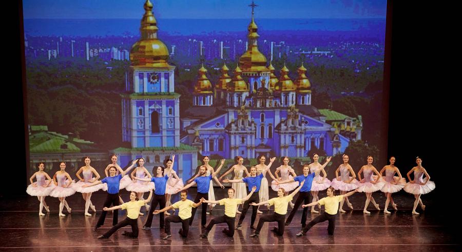 Three rows of dancers stand in front of a colorful, ornate projection of a church. The first row of dancers kneel and wear yellow shirts, the second row behind them stands with blue shirts, and the backmost, third row stands in pink tutus and cream colored dresses.