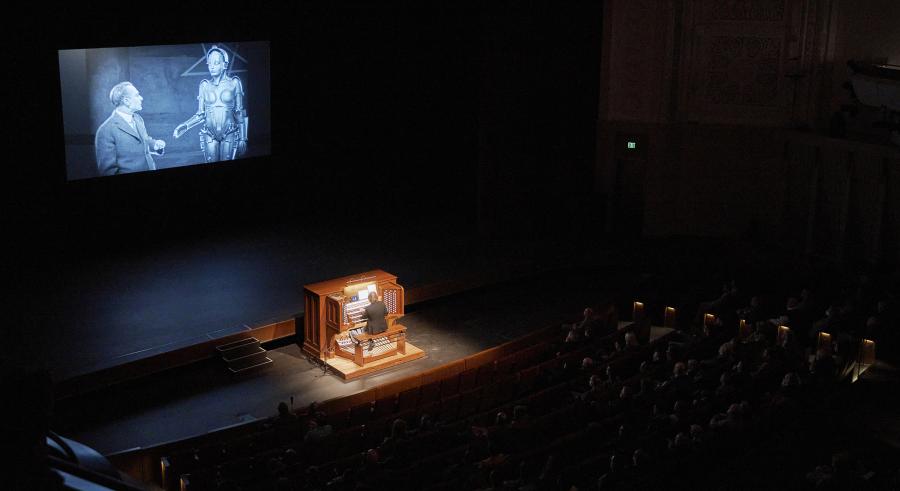 An audience watches a film onstage in a darkly-lit auditorium while an organist plays in the center. 