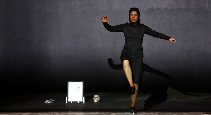 A dancer dressed in an all black, hooded outfit lifts one of their legs with arms outstretched beside them.