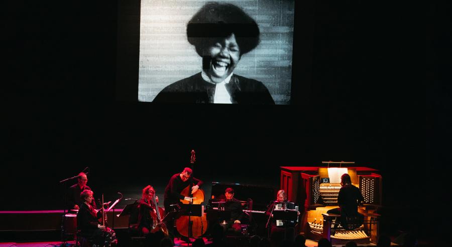 Musicians play on Northrop stage underneath a large black and white photo of a laughing woman from the Cinema’s First Nasty Women film collection.
