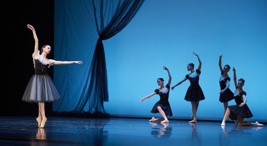Five ballerinas in black and white costumes fill a blue-lit stage. One ballerina stands on point in front of the rest with arms outstretched above and beside their body, while two kneel and two stand with similarly extended arms behind.