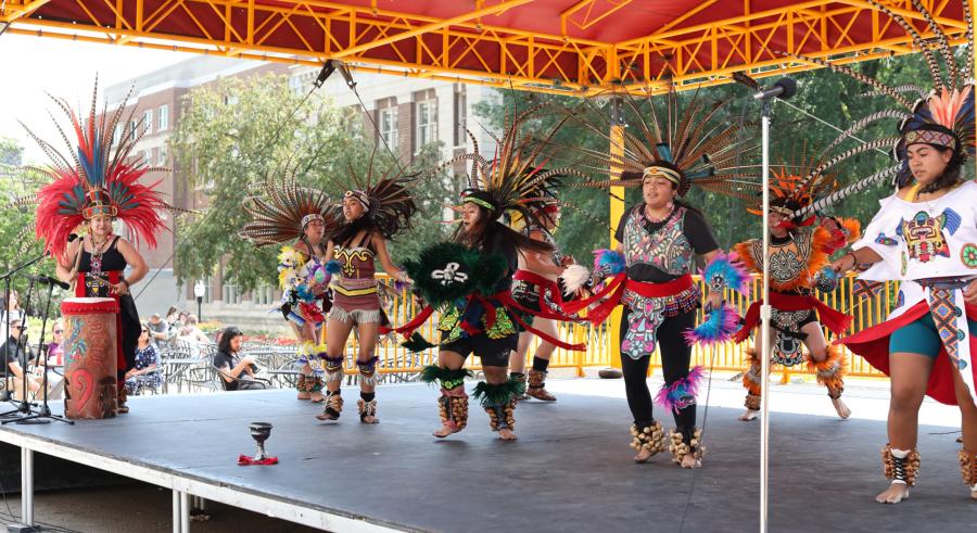 Eight performers from Kalpulli KetzalCoatlicue wearing traditional Aztec and Indigenous dancewear, dancing and playing music on the Northrop Plaza Stage.