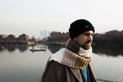 Kit Downes wearing a knit scarf and hat, stands outside by a lake with cityscape in distance.