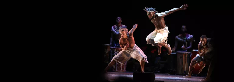 Three dancers wearing white appear on stage in front of musicians playing percussion. One dancer appears in the center, jumping in the air with their arms outstretched behind them.