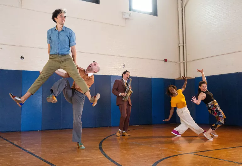 A pair of Swing dancers and a saxophonist playing in the backgound. One couple is lifting their partner in the air