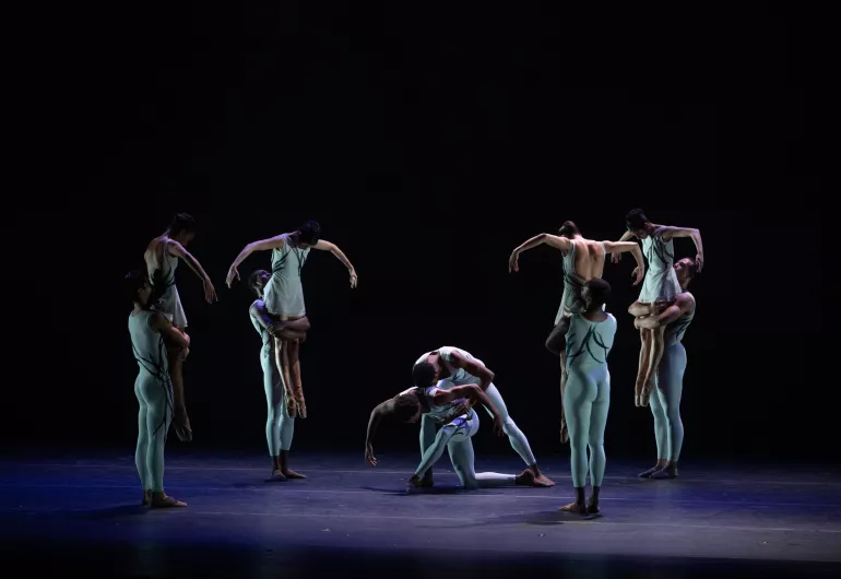 4 female dancers are lifted by their partners, they have their heads down and arms out. They form a circle around another pair in a hug pose at the center.