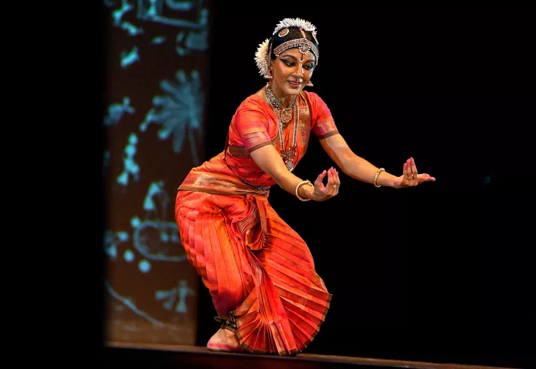 Dancer in red costume leans slight forward, arms in front, fingers pointing up