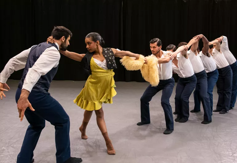 Diagonal line of male dancers, behind female in a yellow dress.