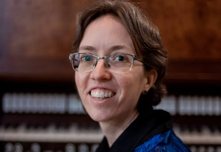 An organist wearing a deep blue button down with short, dark brown hair and glasses sits on a bench in front of a large, wooden organ console with their head turned to the left. 