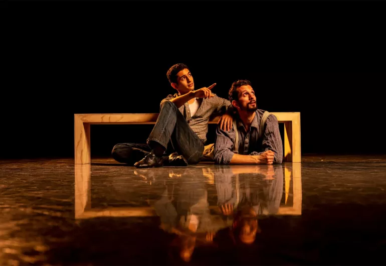 Two male dancers - one casually sitting against and the other lying under a bench. Both looking to the upper right and one is pointing.