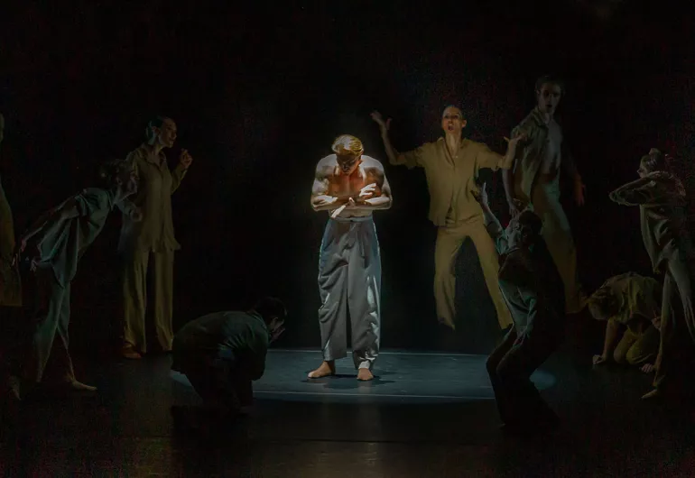 Male dancer in spotlight looks down, dancers surround him with distraught expressions and wild hands