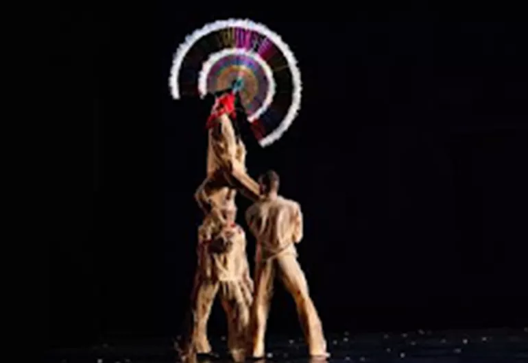 Two dancers hold a third above their shoulders, while a fourth dancers across the stage points a finger at the elevated dancer. The elevated dancer wears a large, circular headpiece, that stands out before the all black background.