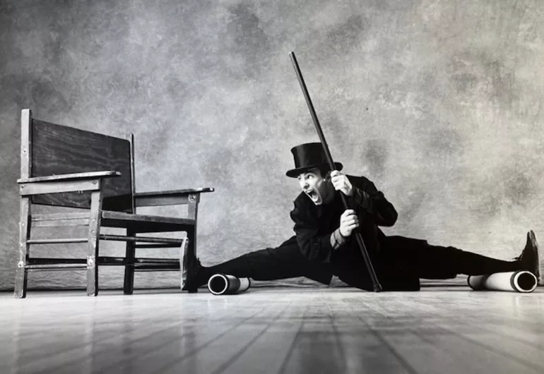 A person's legs are on two cylinders completely spread apart and nearly sitting on the ground. They are wearing a black hat and holding a cane. The person is looking to their right at a chair.