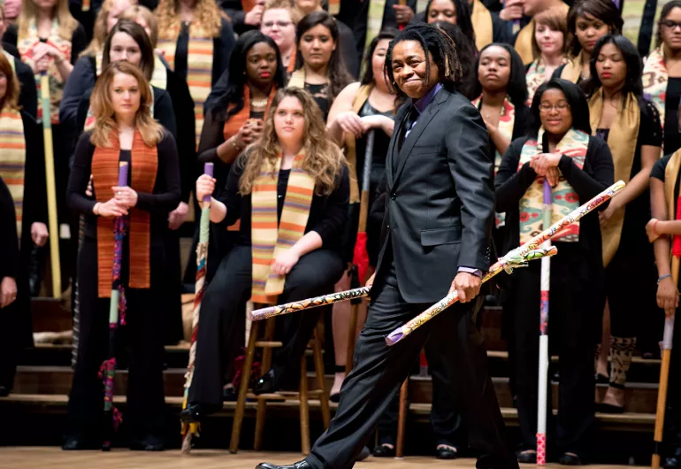 A performer steps out from a choir carrying a colorful stick.