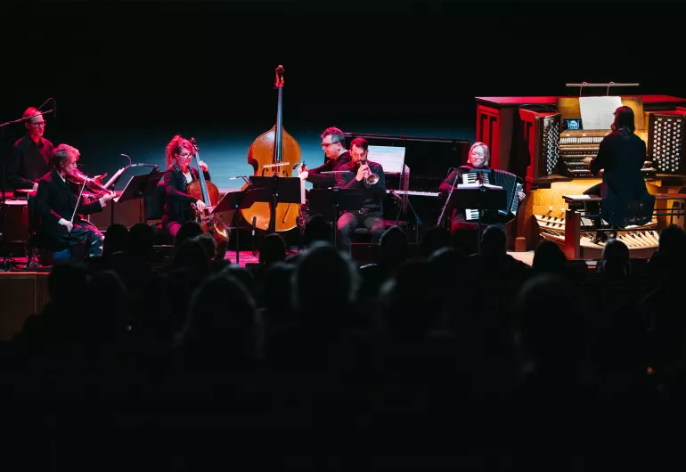 An ensemble of 7 musicians including a drum player, violinist, bassist, cellist, horn player, pianist, accordionist, and pipe organist dressed in black perform onstage with a dark background and pink lighting. 