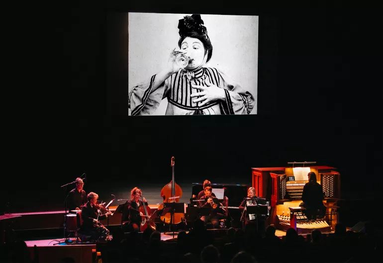 An ensemble of 7 musicians dressed in black perform onstage with a projection of a film above them. The screen shows a black and white film still of a person wearing a striped top, drinking a glass of wine with one hand held to their chest and their eyebrows raised.