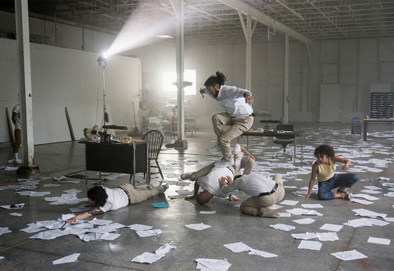 Dancers pose in contorted poses in a large room with scattered papers all over the floor.