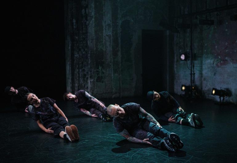 Four dancers dressed in casual dark clothing and sneakers sit on a dark stage with their legs oustretched forward, feet pointing straight up, and slightly leaning on one hip with their bodies pointed to the left.