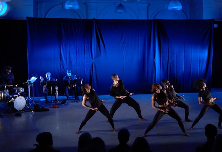 A group of 5 dancers wearing black clothing appear onstage with cobalt blue lighting. 