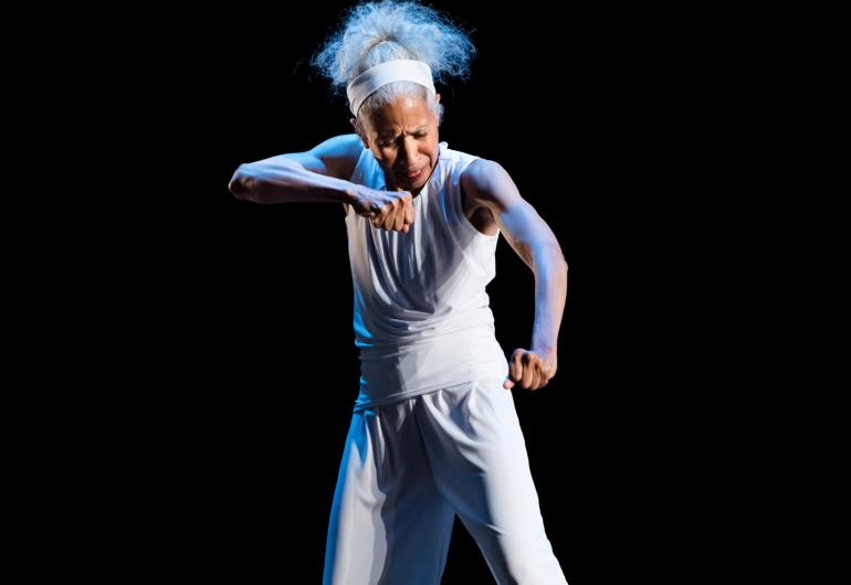 A gray-haired dancer wearing a loose-fitted white tank top and pants stands onstage towards the camera, looking down with one arm raised parallel to the floor and the other slightly lower, their hands clenched.