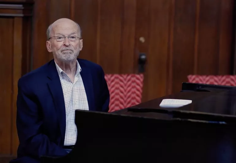 A person in a navy suit and light-colored shirt sit in front of a piano and face the camera. 