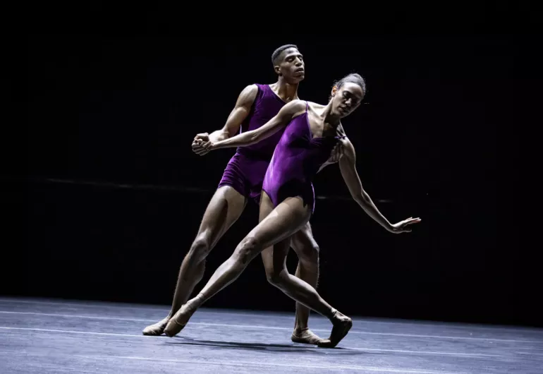 Two Black dancers in purple leotards stand in a graceful dance pose.