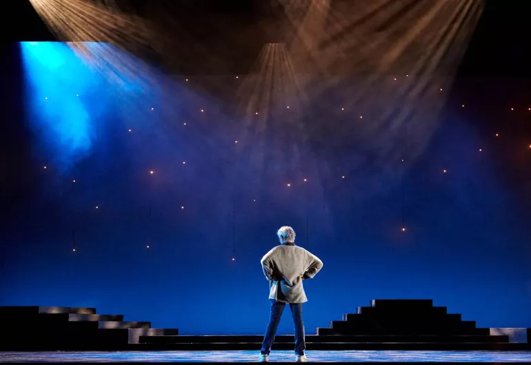 A woman stands on stage with her back turned under spotlights.