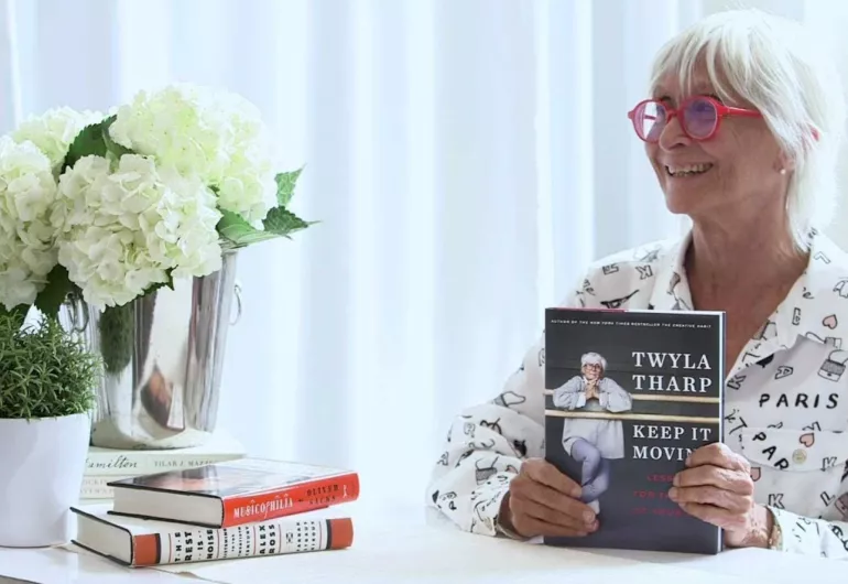 Twyla Tharp with white hair and red glasses, smiles holding her book. The image is linked to YouTube where she is explaining her new book.