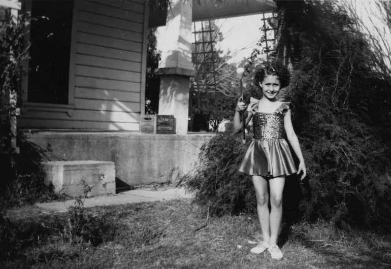 A young, smiling child wearing a dress holds a baton outside a house.