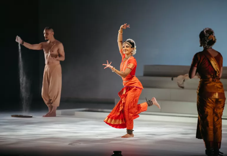 A dancer takes center stage while another stands behind letting sand slowing fall from his hands.