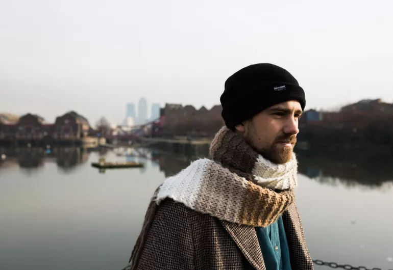 Kit Downes in hat and scarf outside by water with trees and a city skyline in the near distance.