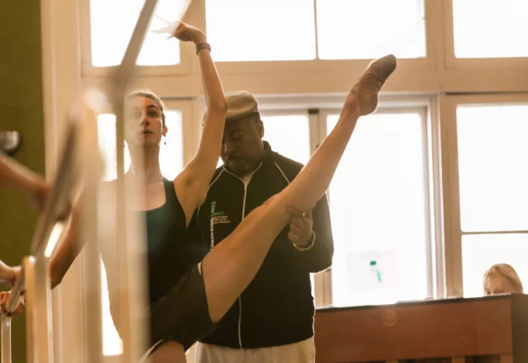 A ballerina stands on pointe with her left leg in the air as Alonzo King stands behind her, guiding her in class. 