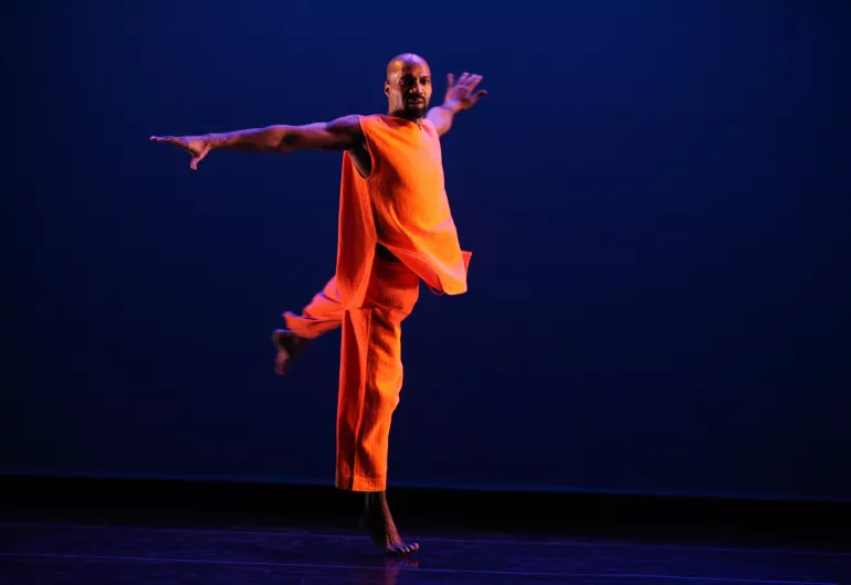 A male dancer dressed in bright orange performs against a purple background.