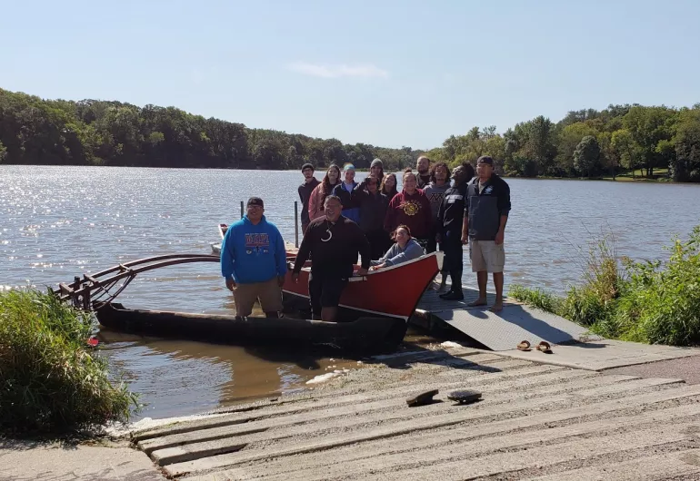 A group of 14 students stand on a dock behind a large canoe floating in a lake.