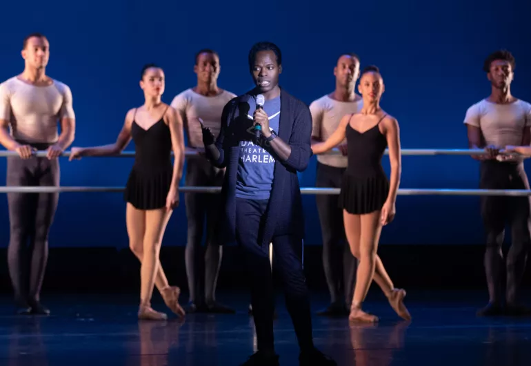 Man stands in the center of the stage holding a microphone in front of dancers at a ballet barre