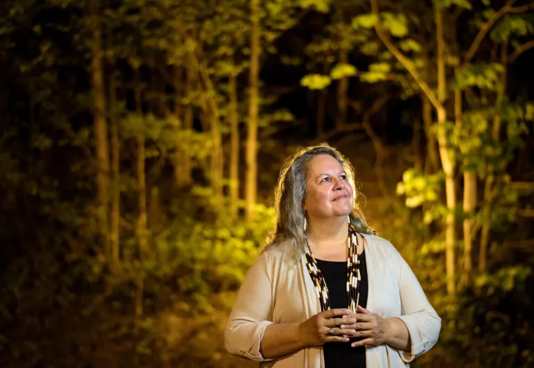 Robin Wall Kimmerer stands among trees looking up to the side smiling