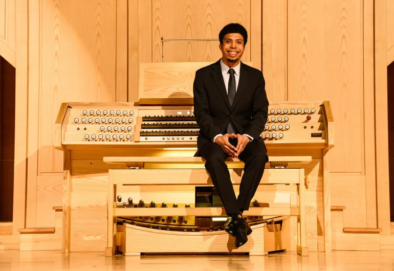 An organist with dark hair wearing a black suit and tie sits in a recital hall with their back towards a wooden organ console and fingers intertwined and placed on their knees.