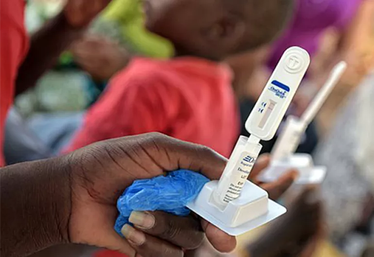 A dark-skinned hand holds up a small white medical device. Out of focus people are shown in the background.