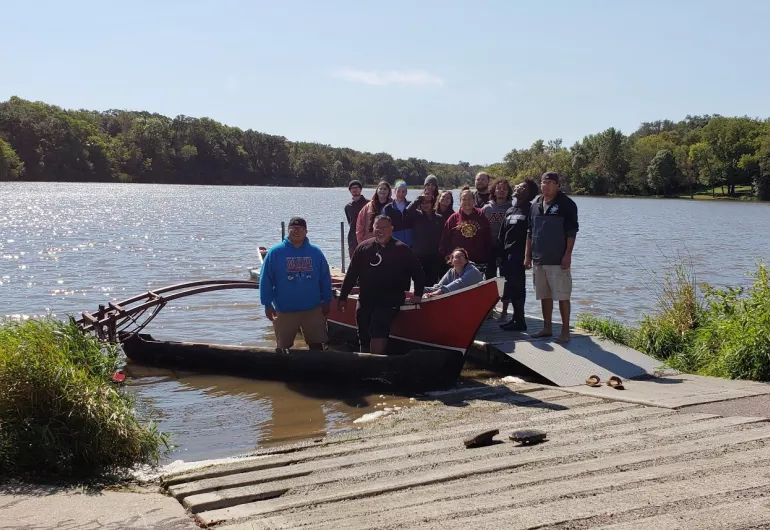 A group of 14 students stand on a dock behind a large canoe floating in a lake.