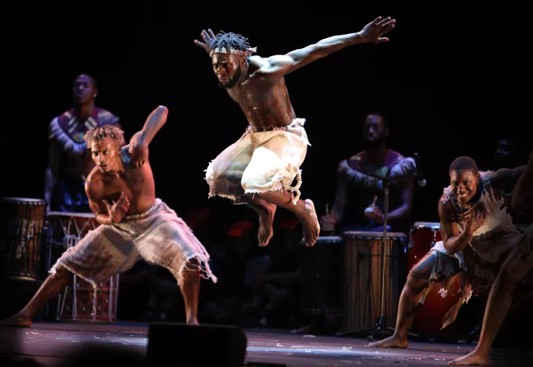 Three dancers wearing white appear on stage in front of musicians playing percussion. One dancer appears in the center, jumping in the air with their arms outstretched behind them.