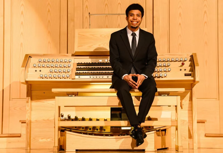 An organist with dark hair wearing a black suit and tie sits in a recital hall with their back towards a wooden organ console and fingers intertwined and placed on their knees.